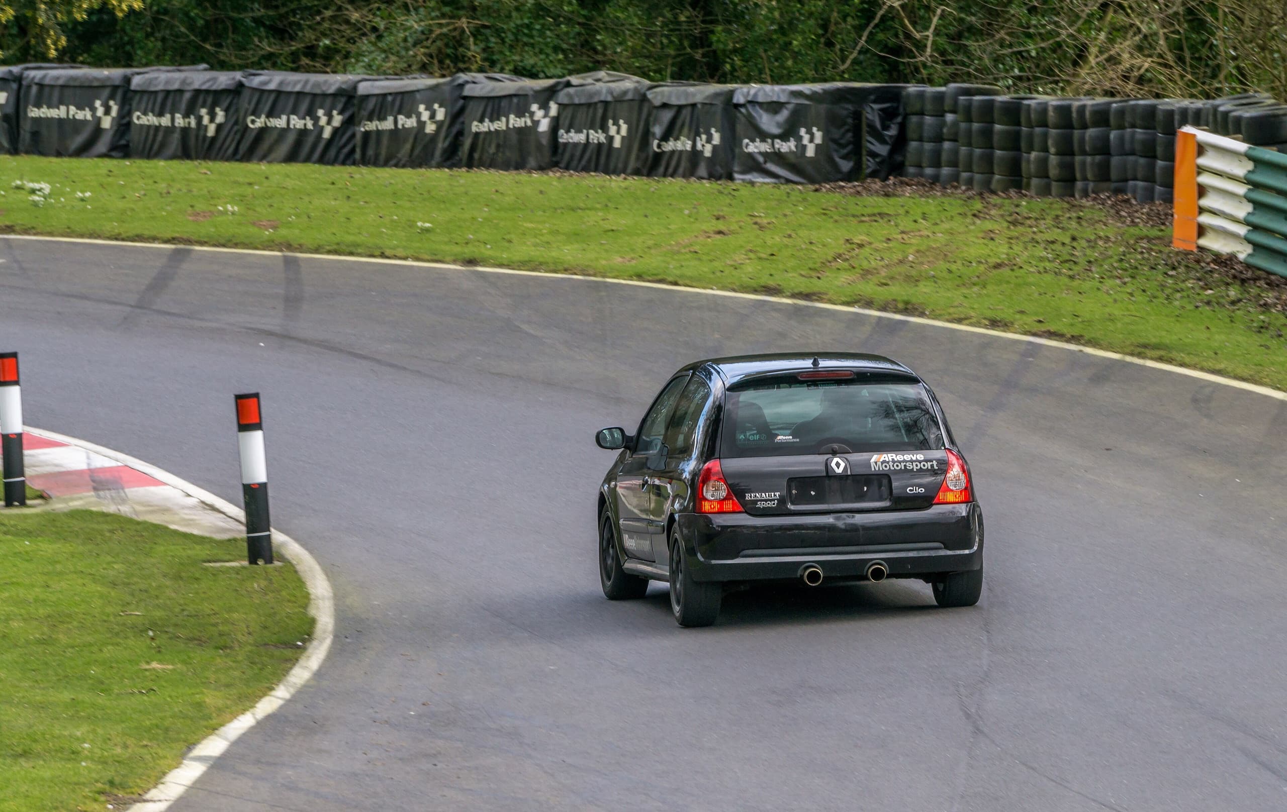 A dark Renault Clio 182 accelerating out of a bend at Cadwell Park, tyre wall behind
